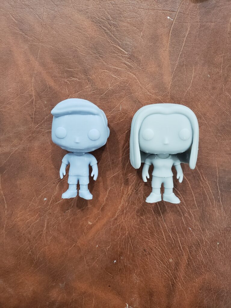 Two custom, gray resin figurines in a Funko-inspired style, featuring large heads, minimal facial details, and 3D printed by PlastiForge3D.