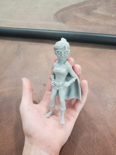 A gray superhero figurine, 3D printed by PlastiForge3D using SLA resin, held in a hand to showcase custom design and high-detail 3D printing services.