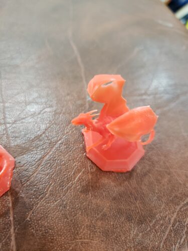 A small, orange dragon miniature, 3D printed by PlastiForge3D for tabletop gaming, showcasing fine detail and SLA resin quality.