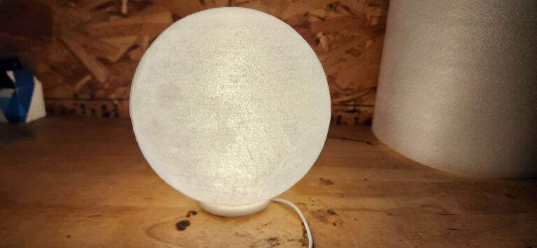 A soft white moon lamp 3D printed from glow-in-the-dark filament, illuminated from within and displaying textured lunar details.