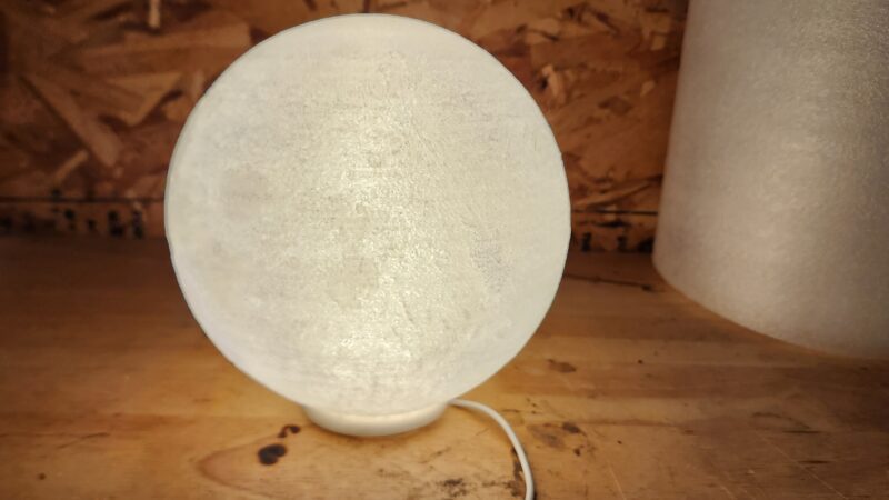 A soft white moon lamp 3D printed from glow-in-the-dark filament, illuminated from within and displaying textured lunar details.