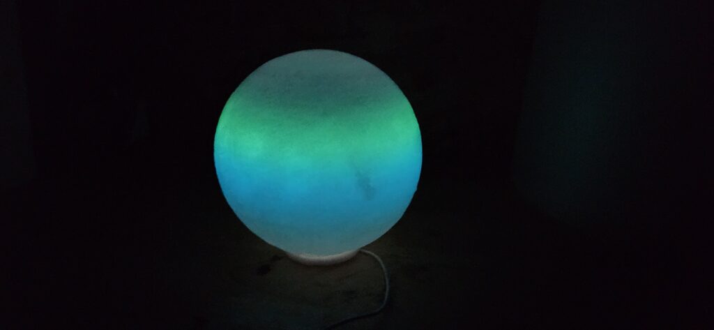 A glowing moon lamp radiating greenish-blue light in a dark room, 3D printed with luminescent filament for a captivating nighttime effect.