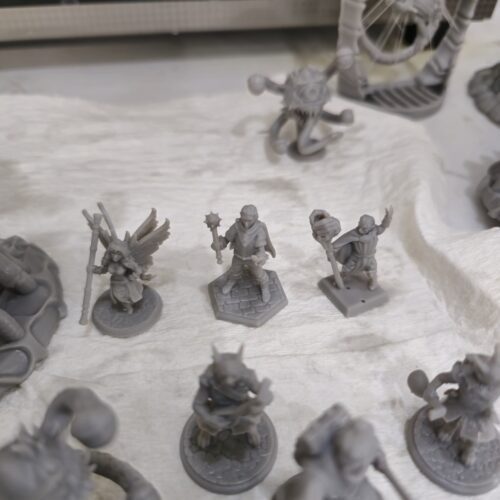 A collection of high-detail tabletop RPG miniatures 3D printed in gray resin by PlastiForge3D, featuring fantasy characters like a wizard, warrior, and fairy.