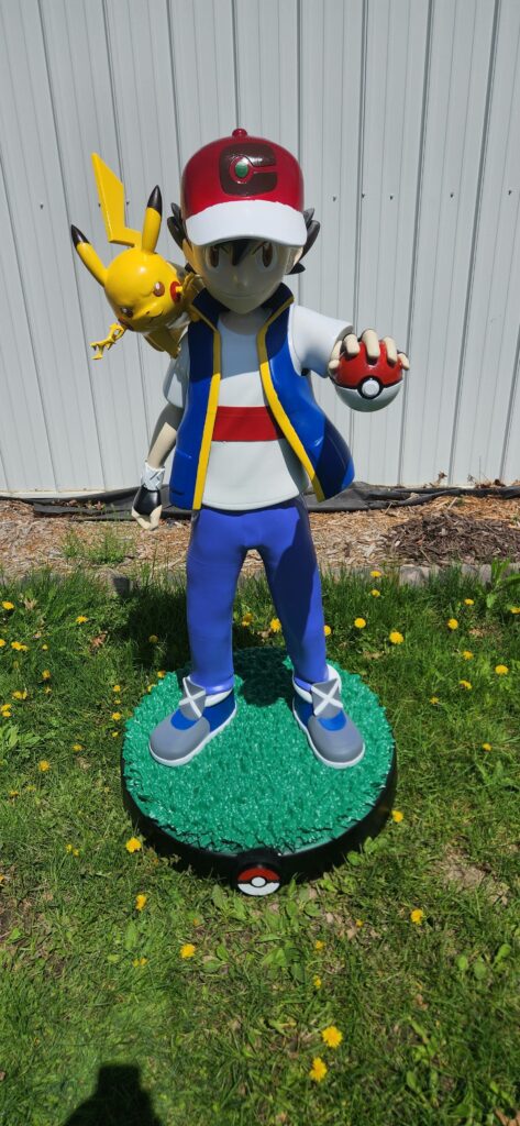 Life size 3D printed and painted statue of Ash Ketchum holding a Pokéball with Pikachu on his shoulder, standing on a custom base outdoors.