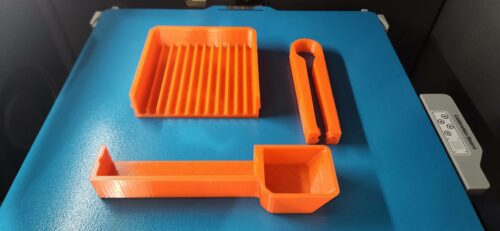 Bright orange PETG tool tray, scoop, and tongs freshly 3D printed on a Bambu Lab textured build surface.