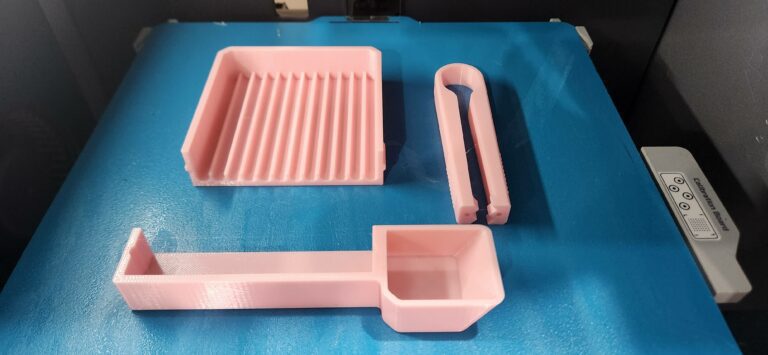 Pastel pink PETG 3D printed tool tray, scoop, and tongs freshly printed on a Bambu Lab textured build plate.